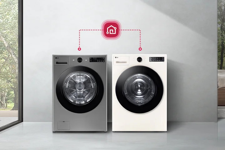 두 대의 LG 세탁기가 나란히 놓여 있으며, 그 위에 스마트홈 아이콘이 연결된 모습이 있습니다. 세탁기 주변으로는 넓은 창문을 통해 나무가 있는 외부 풍경이 보이고, 반대편에는 쿠션이 놓인 소파가 있는 실내 공간이 있습니다.