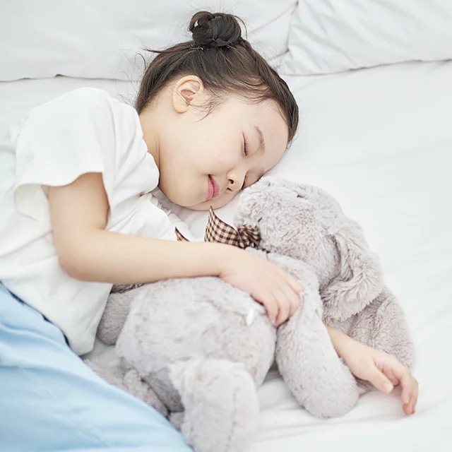 어린아이가 침대에서 회색 곰 인형을 안고 잠들어 있습니다. 아이는 흰색 티셔츠와 파란색 바지를 입고 있으며, 머리는 깔끔하게 묶여 있습니다. 침대는 하얀 시트로 덮여 있습니다.