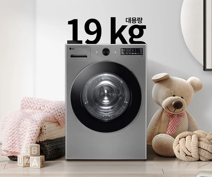세탁기가 놓여 있으며, 상단에 대용량 19kg라는 글자가 있습니다. 세탁기 주변에는 곰 인형과 분홍색 담요가 놓여 있고, 바닥에는 B, U, A가 적힌 나무 블록이 있습니다. 