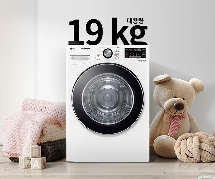 세탁기가 놓여 있으며, 상단에 19kg 대용량이라는 문구가 있습니다. 세탁기 옆에는 곰 인형과 두꺼운 니트 담요가 놓여 있고, 바닥에는 알파벳 블록이 있습니다. 벽에는 추상적인 그림이 걸려 있습니다.