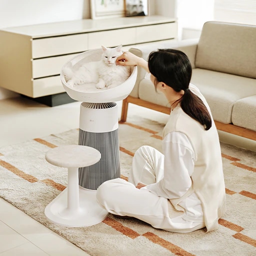 거실에 LG 퓨리케어 AI 오브제컬렉션 에어로캣타워가 배치되어 있습니다. 고양이가 제품의 돔형 좌석에 누워 있고 여성이 러그가 깔린 바닥에 앉아 고양이를 쓰다듬고 있는 모습입니다. 배경에는 서랍장과 페브릭 소파가 있습니다.