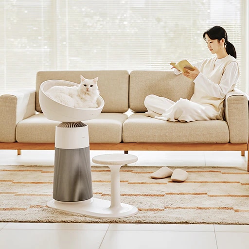 거실에 LG 퓨리케어 AI 오브제컬렉션 에어로캣타워가 배치되어 있습니다. 고양이가 제품의 돔형 좌석에 누워 정면을 바라보고 있습니다. 여성이 페브릭 소파 팔걸이에 기대어 양반다리를 하고 앉아 책을 읽고 있고 소파 아래에는 거실화가 놓여 있습니다. 큰 창문에는 블라인드가 설치되어 있고 바닥에는 러그가 깔려 있습니다.