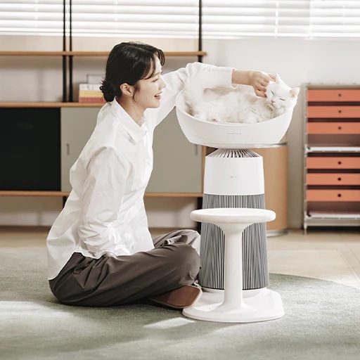 서재에 LG 퓨리케어 AI 오브제컬렉션 에어로캣타워가 배치되어 있습니다. 고양이가 제품의 돔형 좌석에 누워있고 여성이 러그가 깔린 바닥에 앉아 고양이를 쓰다듬고 있는 모습입니다. 배경에는 책장과 주황색의 서류함이 보입니다.