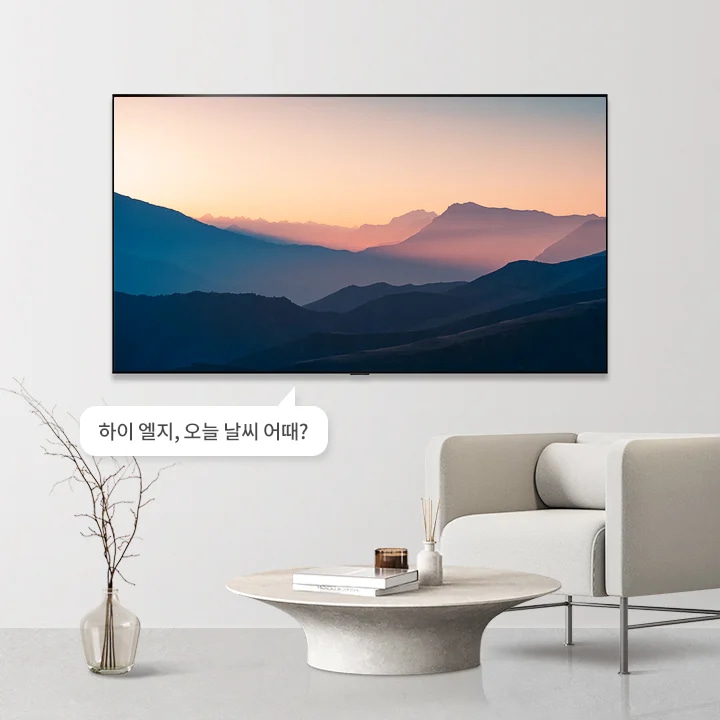 벽에 걸린 대형 화면이 산의 실루엣과 일출 또는 일몰의 장면을 보여주고 있습니다. 화면 아래에는 하이 엘지, 오늘 날씨 어때?라는 텍스트가 포함된 말풍선이 있습니다. 앞에는 둥근 테이블과 그 위에 책과 장식품이 놓여 있으며, 옆에는 베이지색 소파가 배치되어 있습니다. 바닥에는 가지가 꽂힌 투명한 화병이 놓여 있습니다.