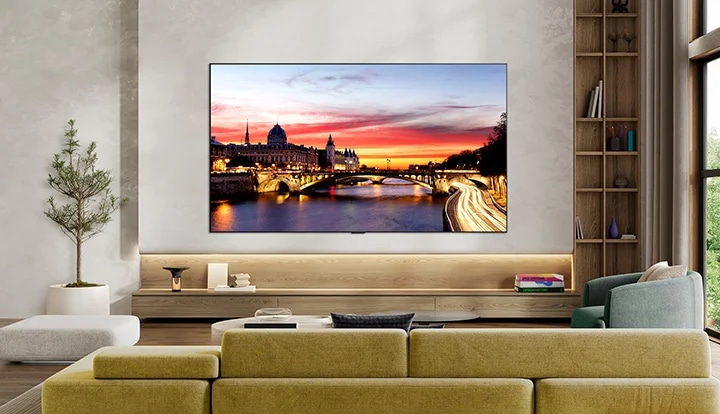 세련된 거실 공간에 벽걸이로 설치되어 있으며, 초슬림 디자인과 얇은 베젤을 갖추고 있습니다. 화면에는 해질녘 강가 도시 풍경이 표시되고 있습니다.
