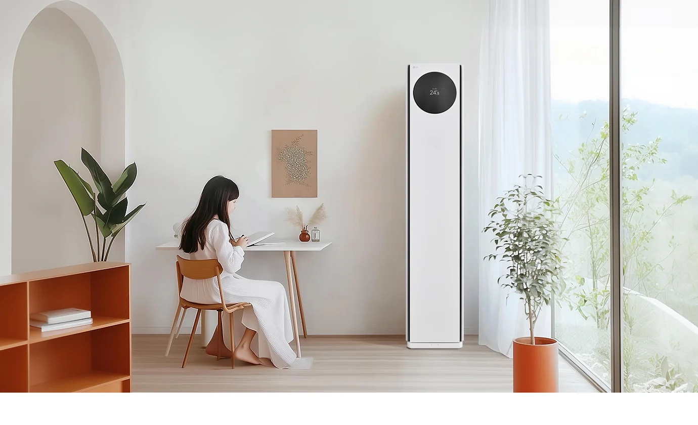 에어컨 상단에는 원형 디스플레이가 있으며, 24.5℃라는 숫자가 표시되어 있습니다. 왼쪽에는 흰색 테이블과 나무 의자에 아이가 앉아 있으며, 테이블 위에는 책과 작은 장식품이 있습니다. 벽에는 식물 액자가 걸려 있으며, 오른쪽에는 커튼이 드리워진 창문과 화분이 보입니다.