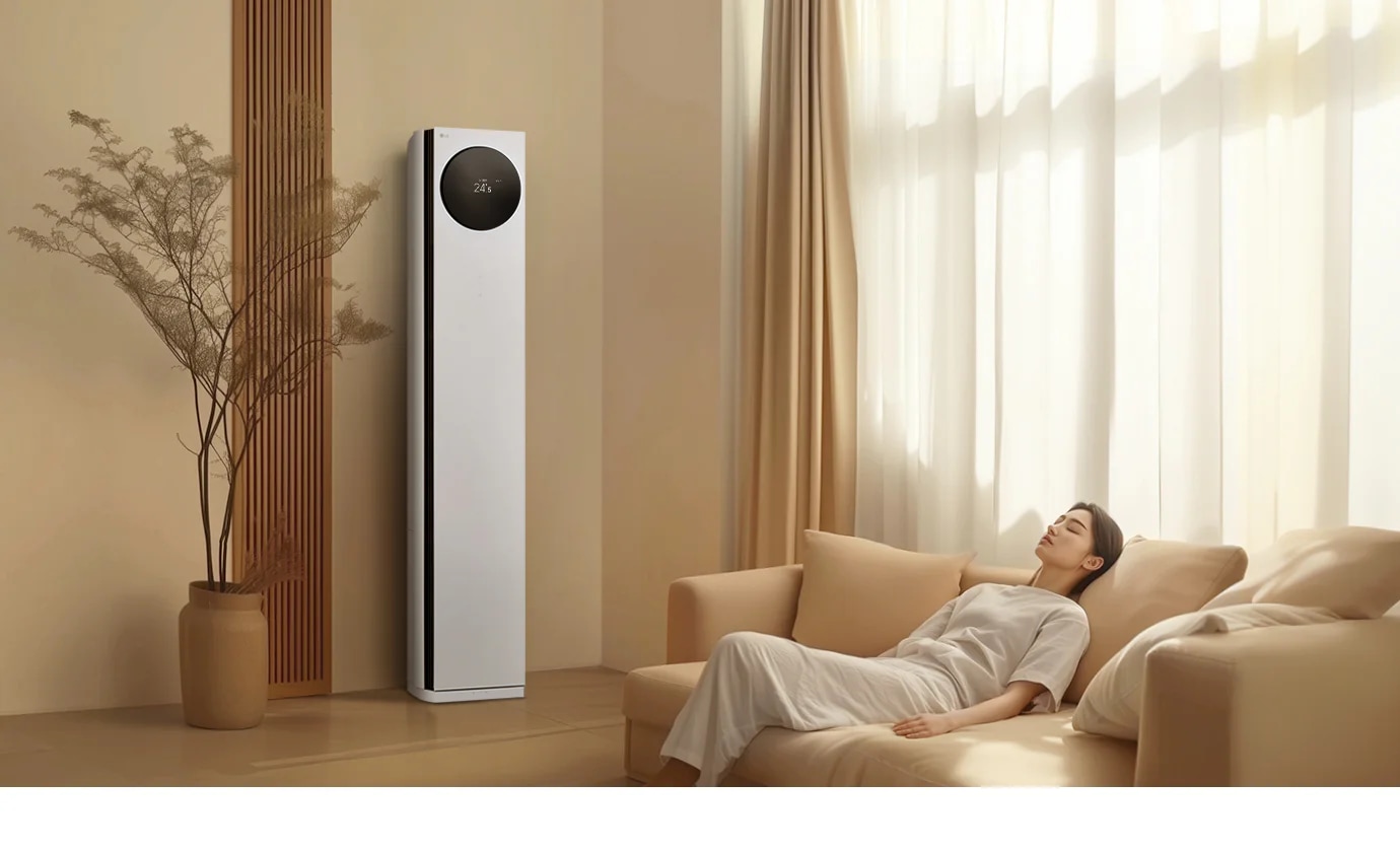 에어컨 상단에는 원형 디스플레이가 있으며, 24.5℃라는 숫자가 표시되어 있습니다. 에어컨 옆에는 나무 소재의 장식 패널과 화분이 놓여 있으며, 오른쪽에는 밝은 색상의 커튼이 드리워진 창문이 있습니다. 창문 앞에는 베이지색 소파가 놓여 있고, 소파에 한 여성이 편안하게 누워 있습니다. 실내는 따뜻한 색조로 꾸며져 있으며, 자연광이 부드럽게 들어오고 있습니다.