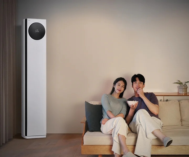 LG 휘센 AI 오브제컬렉션 타워I 에어컨이 왼쪽에 세워져 있으며, 원형 디스플레이에는 24.5℃라는 숫자가 표시되어 있습니다. 오른쪽에는 소파에 앉아 있는 두 사람이 보이며, 남성은 팝콘을 들고 있습니다. 소파 뒤쪽에는 수납장이 놓여 있고 그 위에는 식물과 수건이 놓여 있습니다.