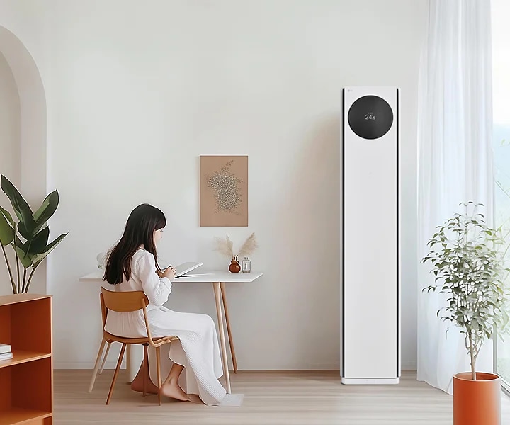 에어컨 상단에는 원형 디스플레이가 있으며, 24.5℃라는 숫자가 표시되어 있습니다. 왼쪽에는 흰색 테이블과 나무 의자에 아이가 앉아 있으며, 테이블 위에는 책과 작은 장식품이 있습니다. 벽에는 식물 액자가 걸려 있으며, 오른쪽에는 커튼이 드리워진 창문과 화분이 보입니다.