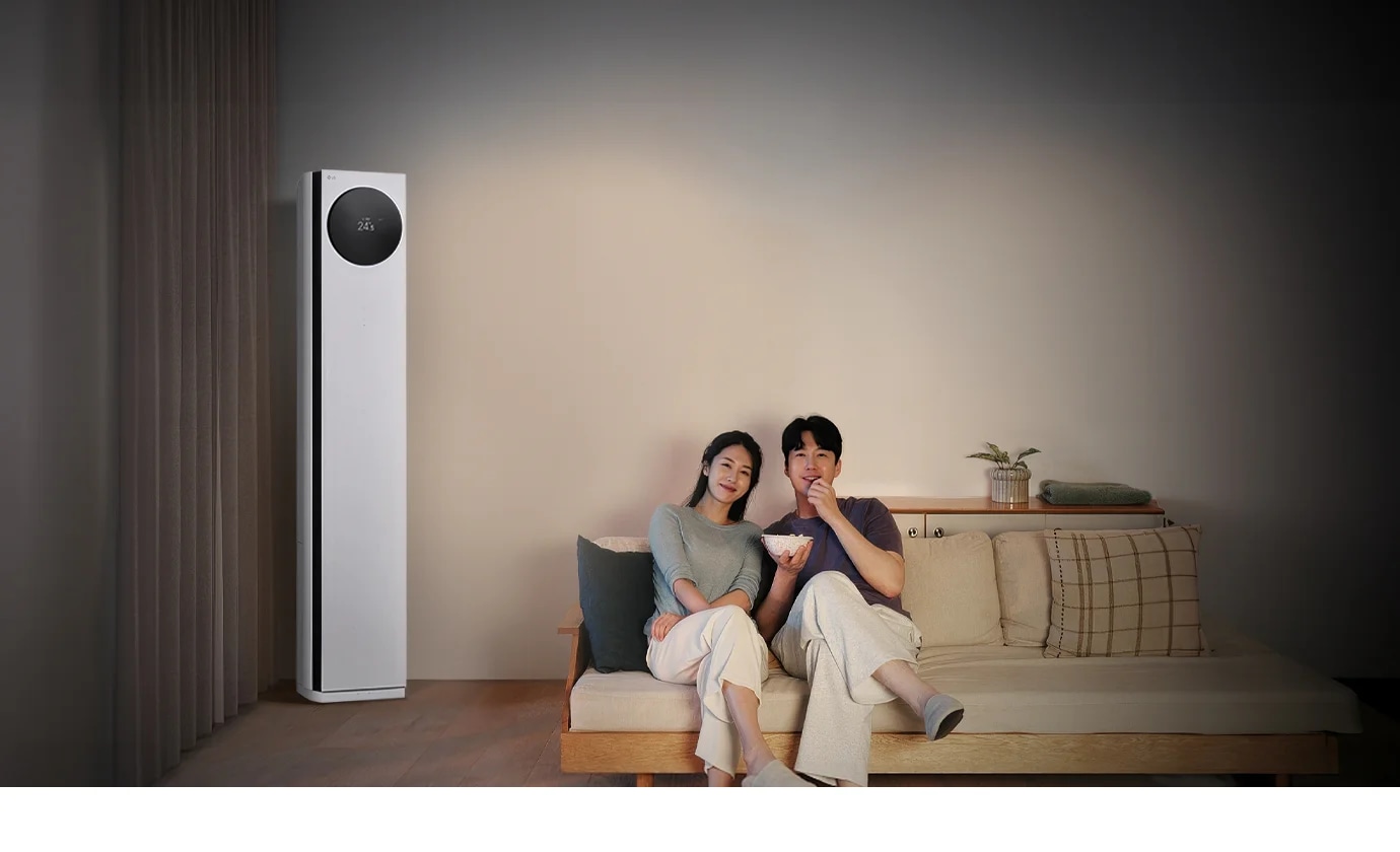 LG 휘센 AI 오브제컬렉션 타워I 에어컨이 왼쪽에 세워져 있으며, 원형 디스플레이에는 24.5℃라는 숫자가 표시되어 있습니다. 오른쪽에는 소파에 앉아 있는 두 사람이 보이며, 남성은 팝콘을 들고 있습니다. 소파 뒤쪽에는 수납장이 놓여 있고 그 위에는 식물과 수건이 놓여 있습니다.