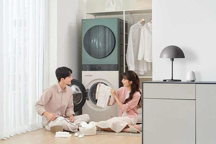 남성과 여성이 세탁기 앞에서 빨래를 정리하며 대화하고 있습니다. 남성은 베이지색 셔츠와 바지를, 여성은 분홍색 상의와 흰색 치마를 입고 있습니다. 세탁기는 두 대가 위아래로 놓여 있으며, 뒤쪽에는 옷걸이에 셔츠가 걸려 있습니다. 바닥에는 빨래 바구니와 접힌 옷들이 놓여 있고, 옆에는 램프가 있는 회색 가구가 보입니다. 창문에는 흰색 커튼이 드리워져 있습니다.
