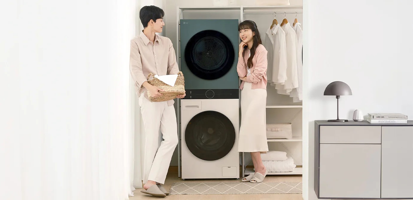 남성과 여성이 세탁기 앞에 서 있습니다. 남성은 빨래 바구니를 들고 있으며, 여성은 세탁기 옆에 기대어 있습니다. 세탁기 위쪽 선반에는 세제와 수건이 놓여 있고, 옷걸이에 셔츠가 걸려 있습니다. 옆에는 램프와 책이 놓인 작은 테이블이 보입니다.