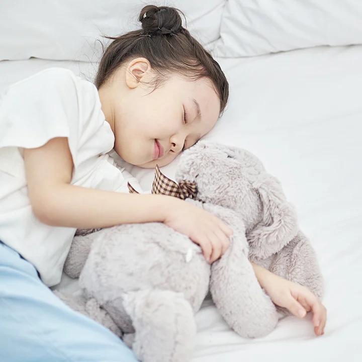 어린아이가 침대에서 회색 곰 인형을 안고 잠들어 있습니다. 아이는 흰색 티셔츠와 파란색 바지를 입고 있으며, 머리는 깔끔하게 묶여 있습니다. 침대는 하얀 시트로 덮여 있습니다.