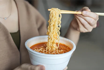 한 사람이 젓가락으로 컵라면을 들고 있으며, 국물이 담긴 흰색 용기에 라면이 담겨 있습니다.