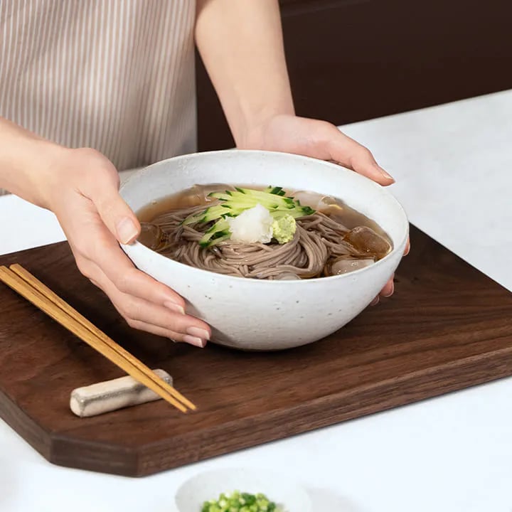 얼음이 들어간 시원한 냉모밀 그릇을 두 손으로 들고 있는 모습입니다. 국수 위에는 채 썬 오이와 무즙 그리고 와사비가 올려져있고 옆에는 나무트레이 위에 젓가락이 놓여있습니다.