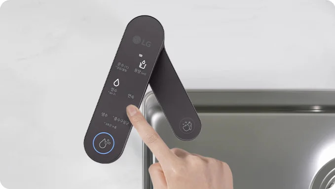 손가락이 LG 로고가 있는 검은색 디지털 패널을 가리키고 있습니다. 패널에는 물 온도, 필터 수명, 정수, 온수, 냉수, 정수추출중이라는 텍스트가 표시되어 있습니다. 패널 옆에는 금속 싱크대가 보입니다.