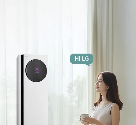 밝은 커튼이 드리워진 실내 공간에서 LG 휘센 AI 오브제컬렉션 타워I 에어컨이 왼쪽에 세워져 있고, 에어컨 본체 상단에 원형 블랙 디지털 디스플레이 화면에 현재 에어컨의 상태를 나타내는 아이콘이 보입니다. 오른쪽에는 컵을 들고 에어컨을 바라보는 여성이 창문을 통해 들어오는 자연광을 받으며 서 있습니다. 여성의 머리 위에는 Hi LG 라는 음성 인식 문구가 표시된 말풍선 형태의 그래픽이 보입니다. 이 이미지는 에어컨의 음성 인식 및 사용자 상호작용 기능을 실내 사용 환경과 함께 보여주고 있습니다.