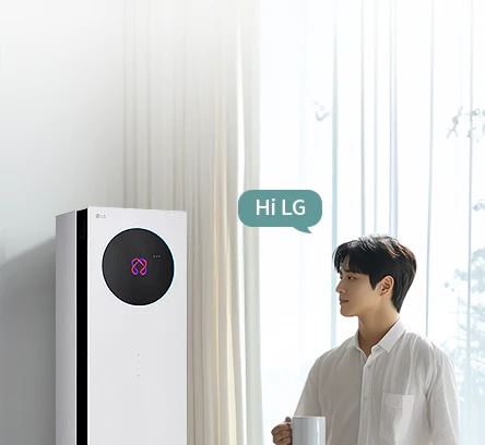 밝은 커튼이 드리워진 실내 공간에서 LG 휘센 AI 오브제컬렉션 뷰I 프로 에어컨이 왼쪽에 세워져 있고, 에어컨 본체 상단에 원형 블랙 디지털 디스플레이 화면에 현재 에어컨의 상태를 나타내는 아이콘이 보입니다. 오른쪽에는 컵을 들고 에어컨을 바라보는 남성이 창문을 통해 들어오는 자연광을 받으며 서 있습니다. 남성의 머리 위에는 Hi LG 라는 음성 인식 문구가 표시된 말풍선 형태의 그래픽이 보입니다. 이 이미지는 에어컨의 음성 인식 및 사용자 상호작용 기능을 실내 사용 환경과 함께 보여주고 있습니다.