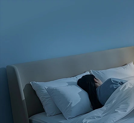 침대에 누워 있는 한 사람이 보이며, 파란색 벽과 베이지색 헤드보드가 있는 침실의 모습입니다. 사람은 회색 잠옷을 입고 흰색 베개와 이불에 둘러싸여 있습니다. 방 안은 조용하고 편안한 분위기를 자아내며, 조명이 은은하게 비추고 있습니다. 에어컨은 이미지에 보이지 않으며, 텍스트도 포함되어 있지 않습니다.