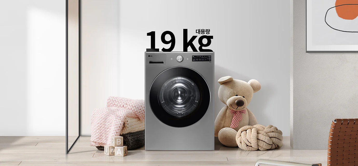 세탁기가 놓여 있으며, 그 위에 19kg 대용량이라는 문구가 적혀 있습니다. 세탁기 주변에는 분홍색 니트 담요와 곰 인형이 놓여 있고, 바닥에는 알파벳 블록이 있습니다. 