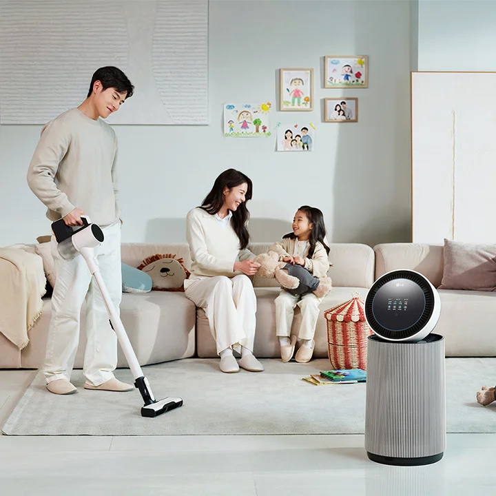 거실 공간에 LG 퓨리케어 AI 오브제컬렉션 360˚ 공기청정기가 있습니다. 소파에 앉아 있는 남성과 뛰어노는 아이, 그리고 청소기를 들고 있는 여성이 보입니다. 벽에는 그림이 걸려 있고, 바닥에는 화분과 장난감 등이 배치되어 있습니다.