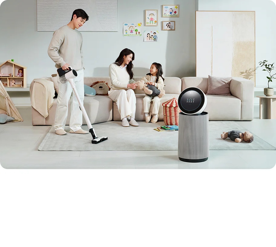 거실 공간에 LG 퓨리케어 AI 오브제컬렉션 360˚ 공기청정기가 있습니다. 소파에 앉아 있는 남성과 뛰어노는 아이, 그리고 청소기를 들고 있는 여성이 보입니다. 벽에는 그림이 걸려 있고, 바닥에는 화분과 장난감 등이 배치되어 있습니다.