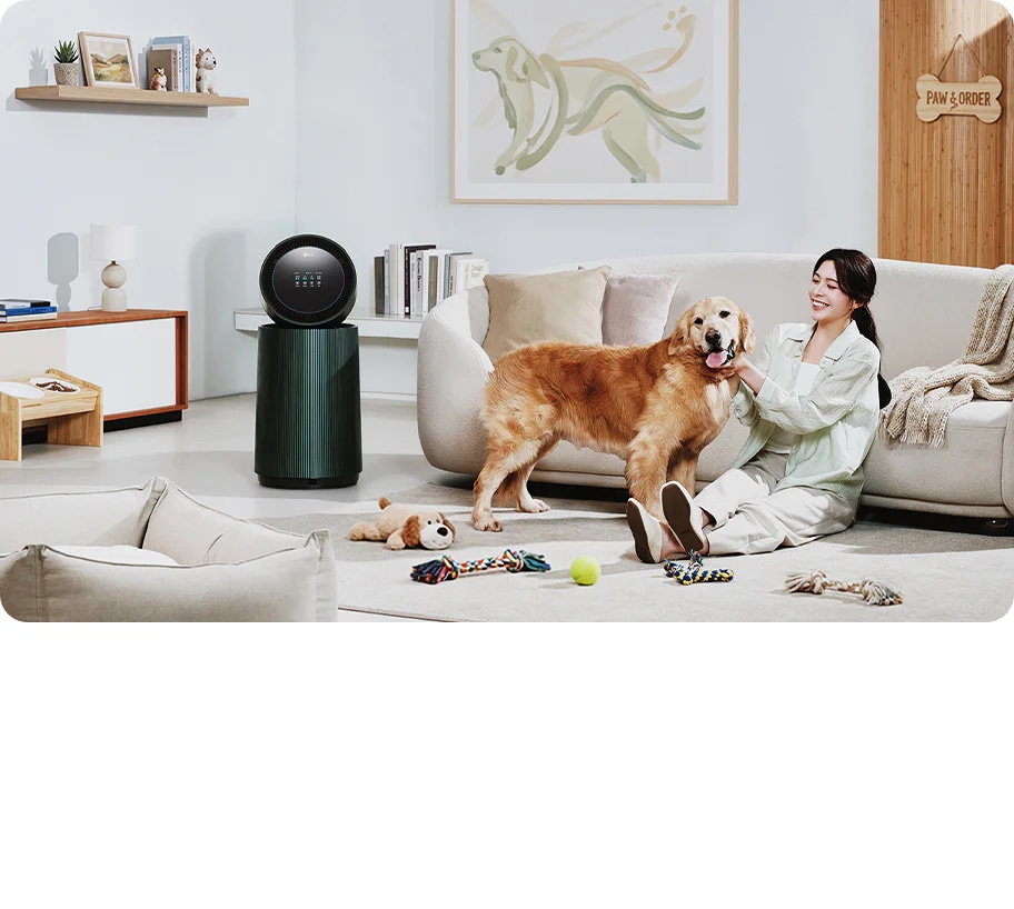 거실 공간에 LG 퓨리케어 AI 오브제컬렉션 360˚ 공기청정기가 놓여 있습니다. 한 여성이 앉아있는 강아지를 만지고 있으며 주변에는 강아지 장난감이 있습니다. 소파가 보이며, 벽에는 강아지 그림이 걸려 있고 선반에는 책과 장식품들이 놓여 있습니다.
