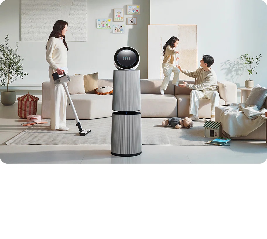 거실 공간에 LG 퓨리케어 AI 오브제컬렉션 360˚ 공기청정기가 있습니다. 소파에 앉아 있는 남성과 뛰어노는 아이, 그리고 청소기를 들고 있는 여성이 보입니다. 벽에는 그림이 걸려 있고, 바닥에는 화분과 장난감 등이 배치되어 있습니다.