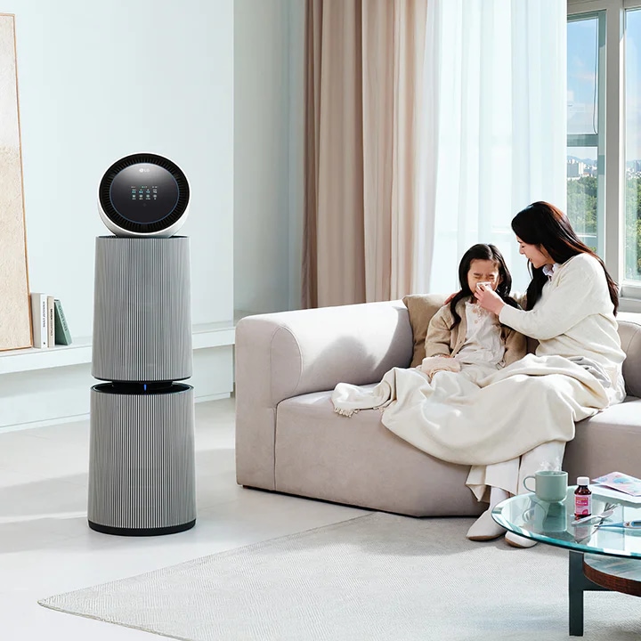 거실 공간에 LG 퓨리케어 AI 오브제컬렉션 360˚ 공기청정기가 놓여 있습니다. 소파 위에는 두 사람이 담요를 덮고 앉아 있으며, 한 여성이 아이의 코를 풀어주고 있는 모습입니다. 테이블 위에는 컵, 약봉투, 약이 놓여 있습니다.